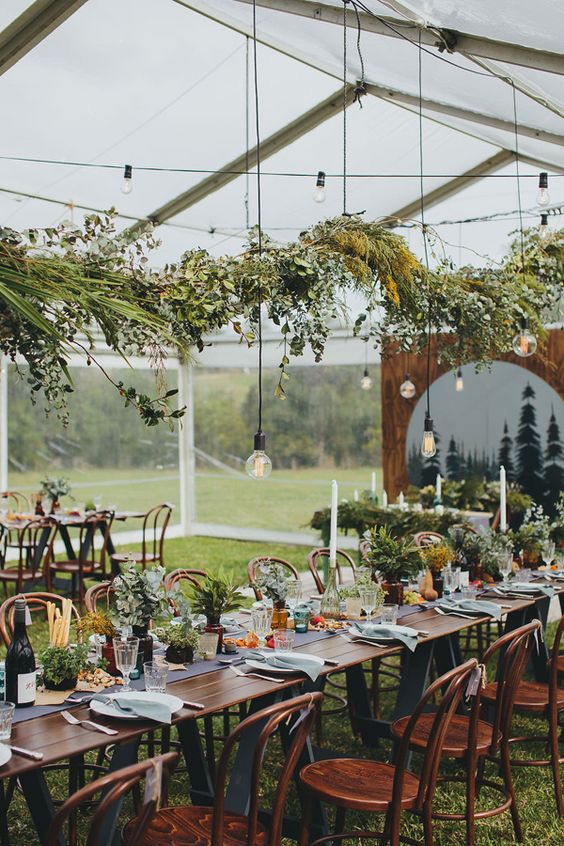 a casual wedding tablescape with potted plants and candles, greenery over the table and some bulbs hanging down