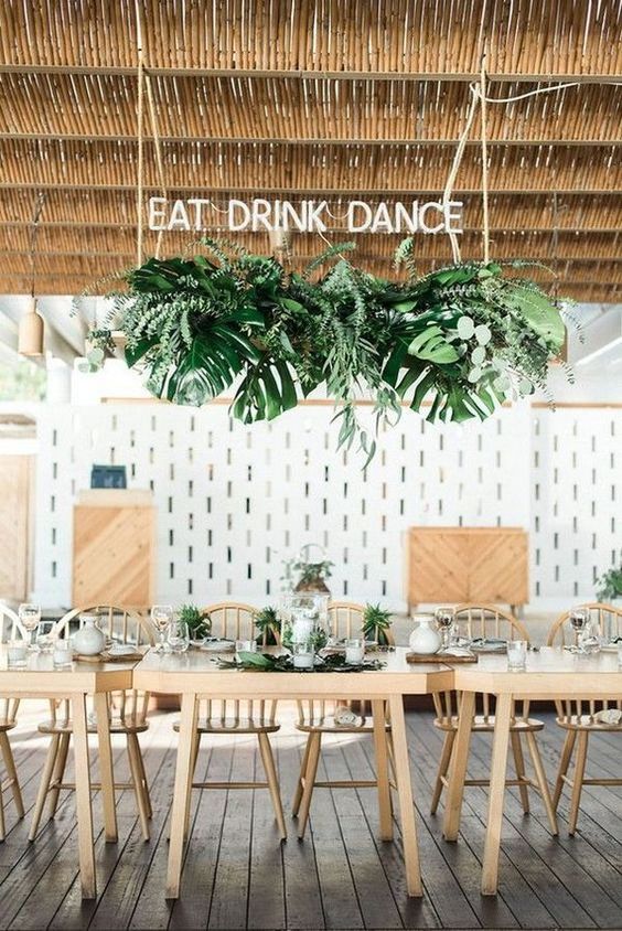 a chic modern tropical wedding tablescape with greenery on the table, a leaf and frond overhead installation, a neon sign