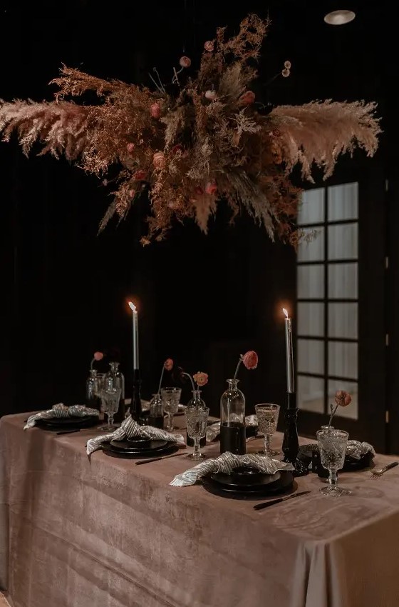 a chic moody rock wedding tablescape with a dusty pink tablecloth, black plates, black vases and candleholders, a grass and bloom overhead isntallation