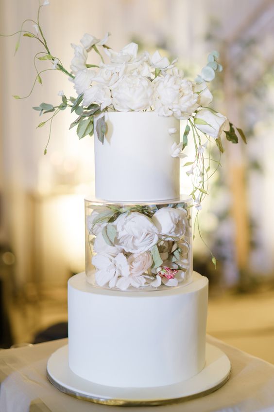 a classic white wedding cake spruced up with a clear tier, white blooms and greenery inside and white blooms on top