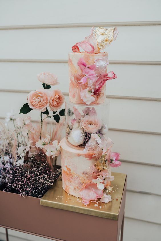 a colorful wedding cake with pink and gold swirls and a clear acrylic separator and pink blooms and gilded branches inside
