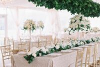 a glam wedding tablescape with a copper tablecloth, white blooms and greenery on the table and a greenery installation over the table
