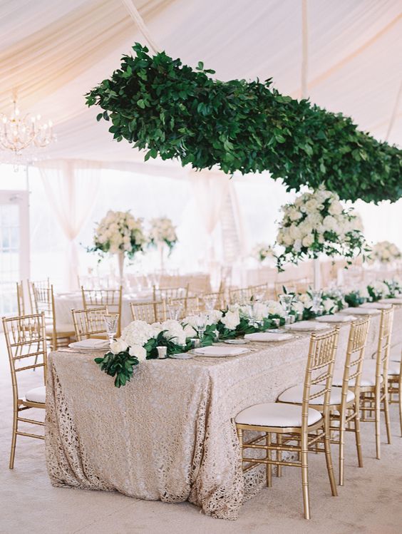 a glam wedding tablescape with a copper tablecloth, white blooms and greenery on the table and a greenery installation over the table