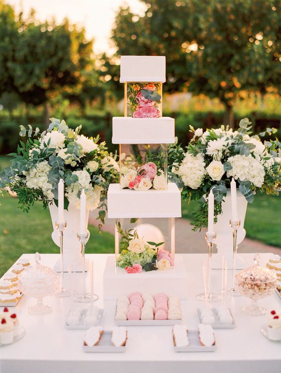 a gorgeous white square wedding cake with clear acrylic separators filled with neutral and bright blooms and greenery is amazing for a garden wedding