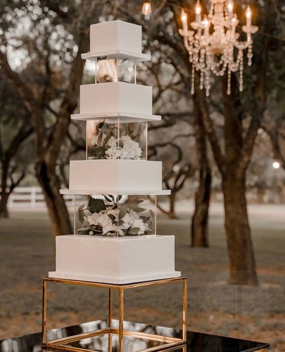 a modern square white wedding cake with clear acrylic separators filled with white blooms and greenery is a stylish idea for a refined wedding
