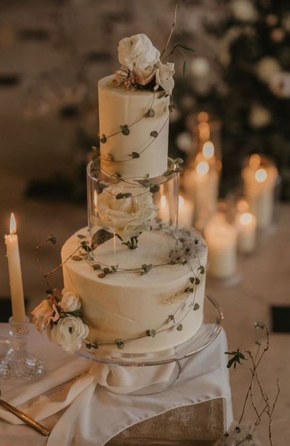 a neutral round wedding cake with a clear stand with a white flower inside, some white blooms and greenery on top is a chic idea
