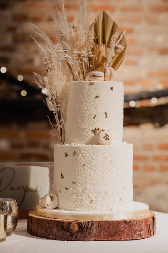 a neutral textural and patterned wedding cake with dried blooms, macarons, beads, dried grasses and a frond is great for a boho wedding