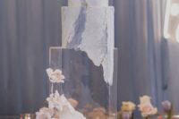 a quirky square wedding cake in blue and white, with much texture and watercolors, with a clear tier and some white and blush blooms