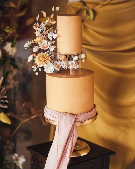 a sophisticated rust colored wedding cake with a clear acrylic tier decorated with dried and fresh blooms and leaves, with a pink ribbon