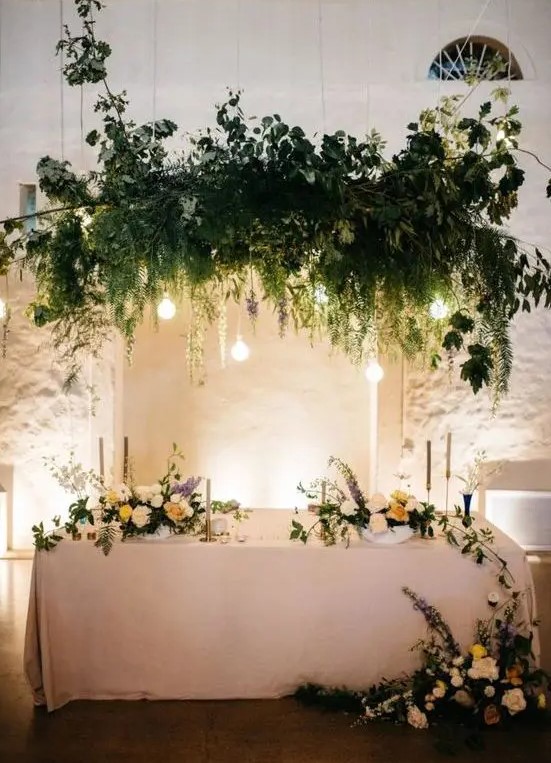a sophisticated wedding sweetheart table with a greenery and bulb overhead installation and refined blooms on and around the table