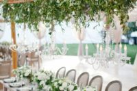 a sophisticated wedding tablescape with neutral linens, white blooms and greenery, an overhead greenery installation and chandeliers