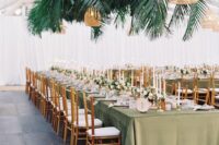 a tropical wedding reception space wiht a green tablecloth, tall and thin candles, fronds and leaves plus woven lamps over the table