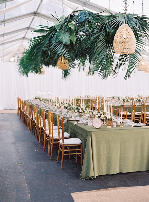 a tropical wedding reception space wiht a green tablecloth, tall and thin candles, fronds and leaves plus woven lamps over the table