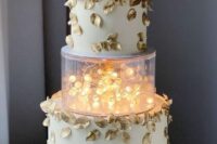 a whimsical wedding cake in white, with gold leaves and a clear acrylic separator with lights plus a gold monogram topper