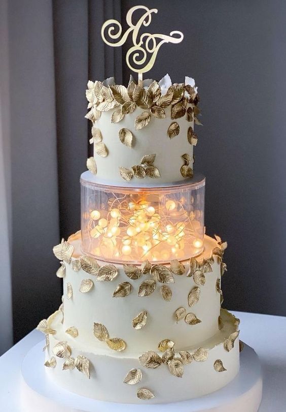a whimsical wedding cake in white, with gold leaves and a clear acrylic separator with lights plus a gold monogram topper