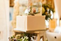 a white textural square wedding cake with a clear acrylic stand with blooms and greenery is a whisical idea for a wedding