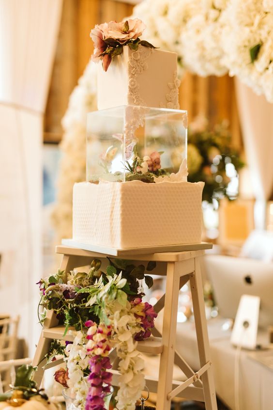 a white textural square wedding cake with a clear acrylic stand with blooms and greenery is a whisical idea for a wedding