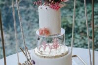 a white textural wedding cake with a clear acrylic stand with pink blooms there and a frond plus dried touches on top is wow