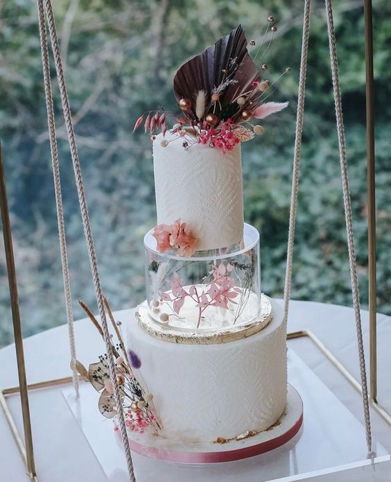 a white textural wedding cake with a clear acrylic stand with pink blooms there and a frond plus dried touches on top is wow