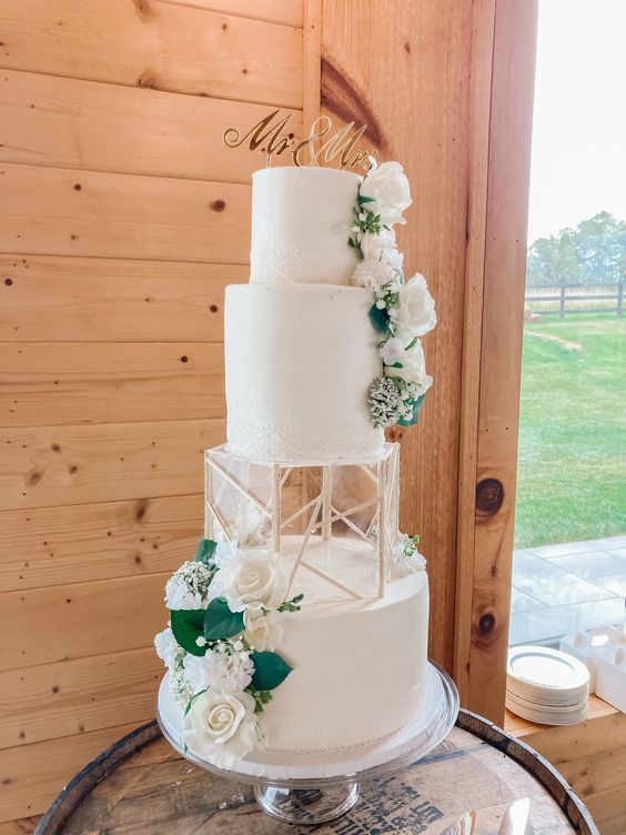 a white wedding cake with an acrylic separator with geometric decor, white blooms and greenery and a gold calligraphy topper