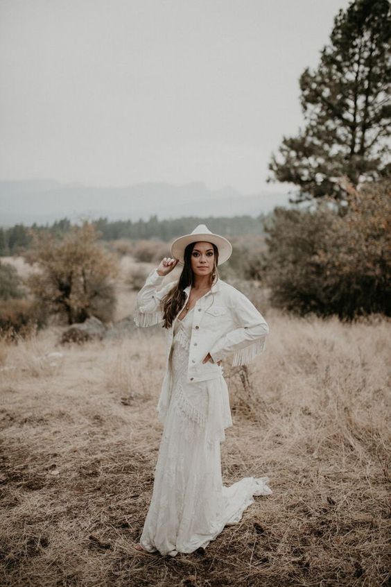 a white denim bridal jacket with fringe on the sleeves, a boho lace wedding dress and a white hat for a boho bride