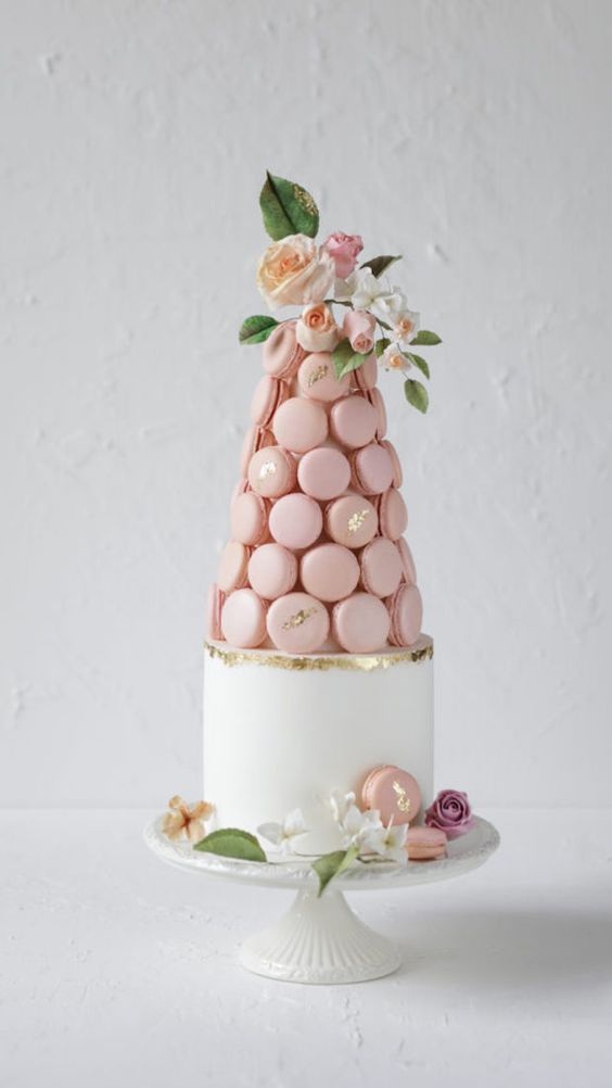 a white wedding cake with a gold foil touch and a pink macaron tower on top, with blusha nd peachy blooms and greenery