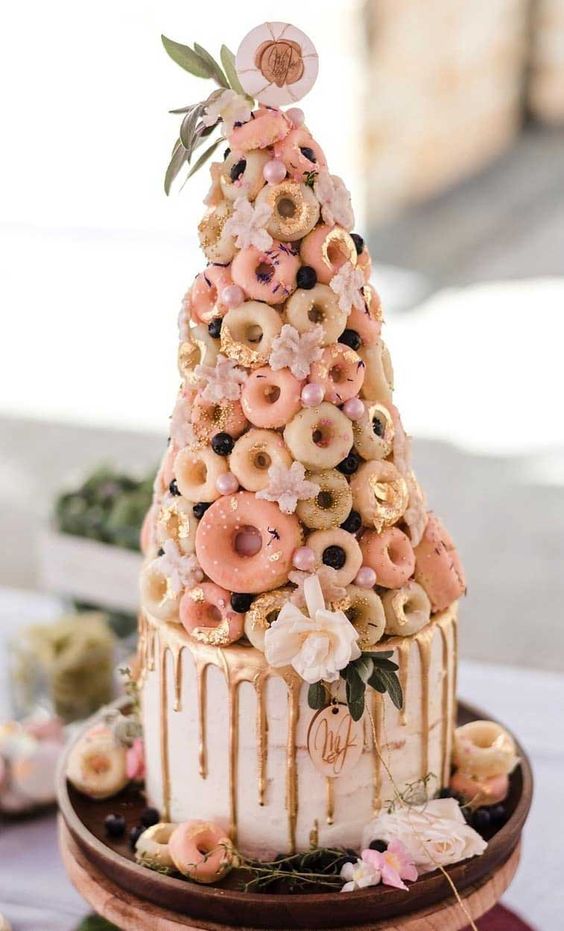 a whimsy pink wedding cake with gold drip and a colorful glazed donut tower, with gold foil, pink beads and greenery