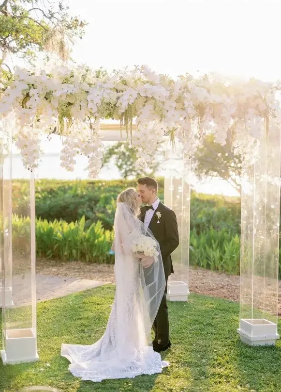 a large acrylic wedding arbor covered with lush white orchids and with a view of a field as a backdrop