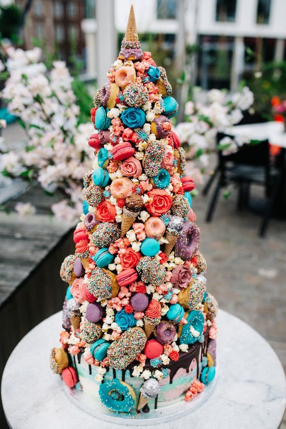 a crazy and super colorful wedding cake topped with a croquembouche of turquoise and coral macarons, glazed donuts, ice cream cones and candies is wow