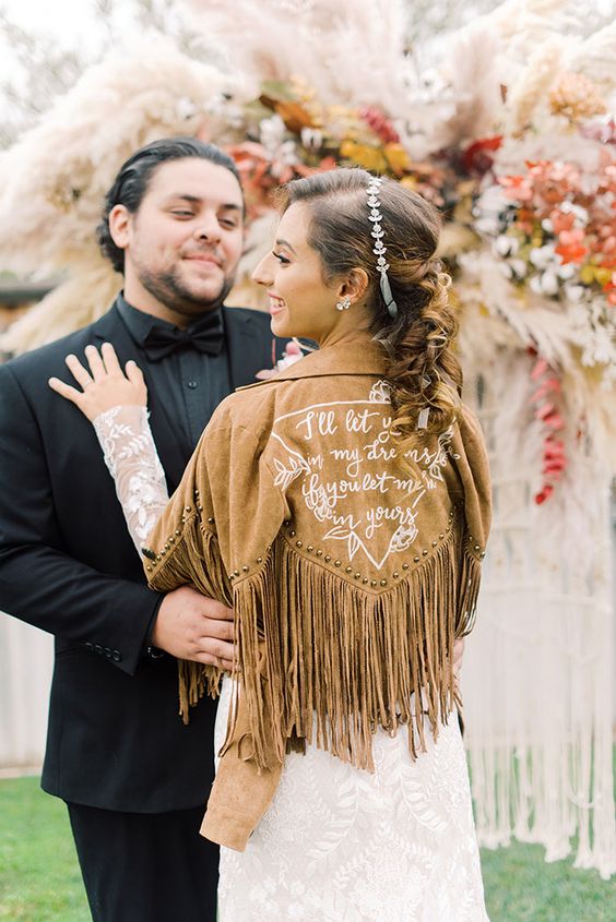 a tan suede bridal jacket with white calligraphy and sutds plus long fringe is a perfect addition to a boho bridal look
