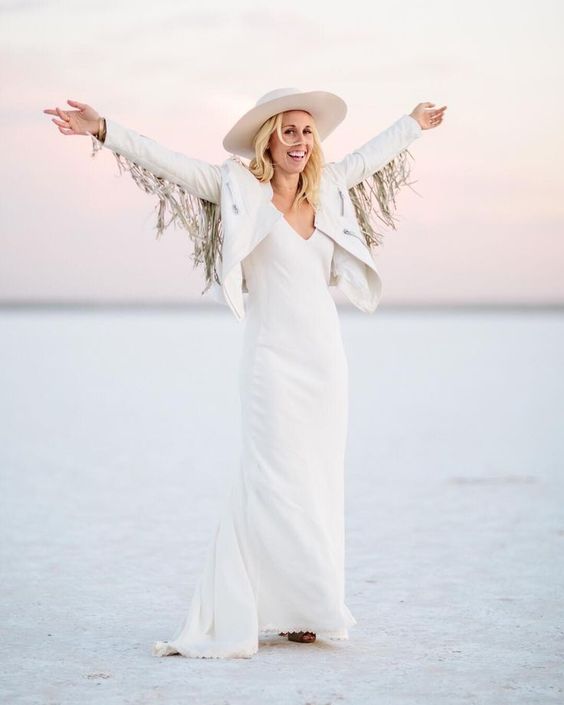 a boho bridal outfit with a plain white wedding dress, a creamy hat, a white jacket with fringe on the sleeves