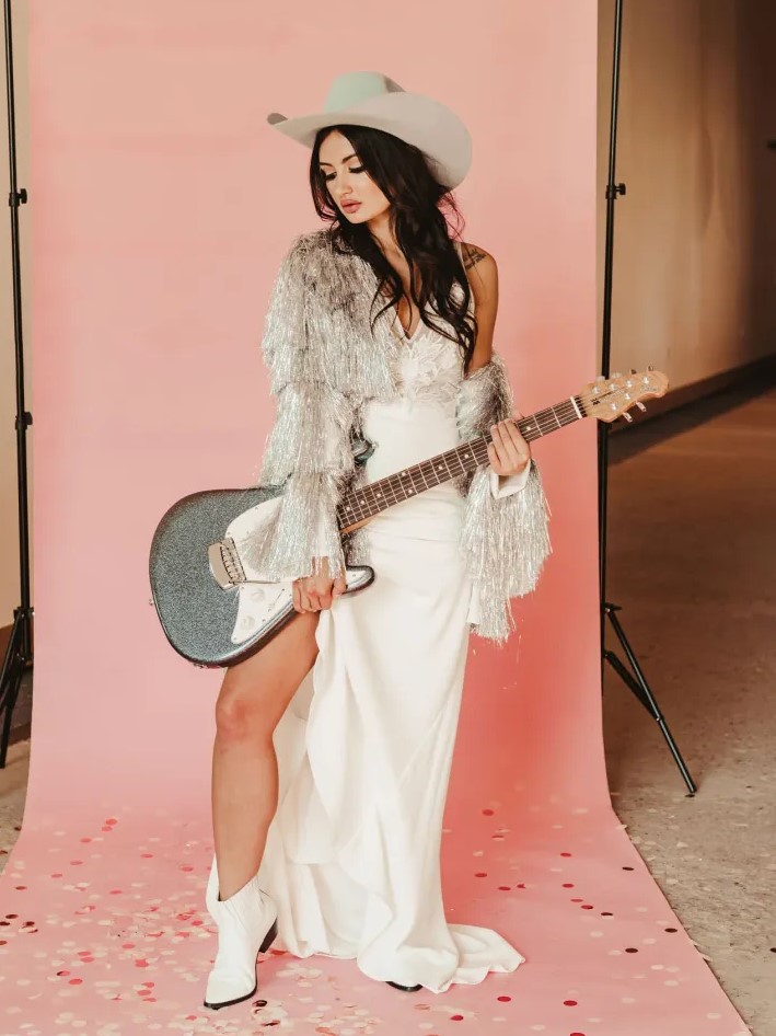 a boho bridal look with a plain wedding dress, a silver fringe jacket, white cowboy boots and a white hat is wow
