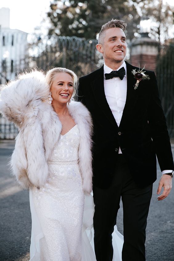 a glam shiny wedding dress paired with a white faux fur jacket and glam flower earrings are an amazing glam bridal look