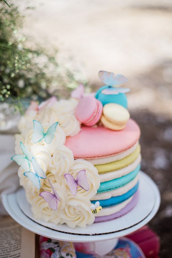 a rainbow macaron wedding cake with vanilla cream, vanilla roses and pastel butterflies and macarons on top is amazing