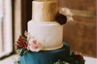 a stylish wedding cake with a gold, two white and teal tiers, white, blush and burgundy blooms and greenery plus a calligraphy topper