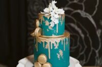 a teal wedding cake with gold drip, gold leaf touches, gold macarons and white blooms on top the cake