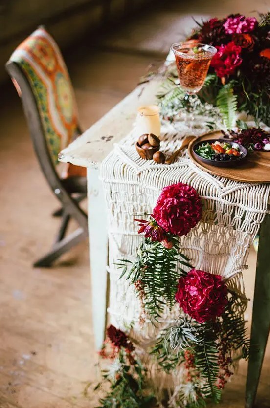a macrame table runner with crimson blooms and greenery for a boho fall wedding
