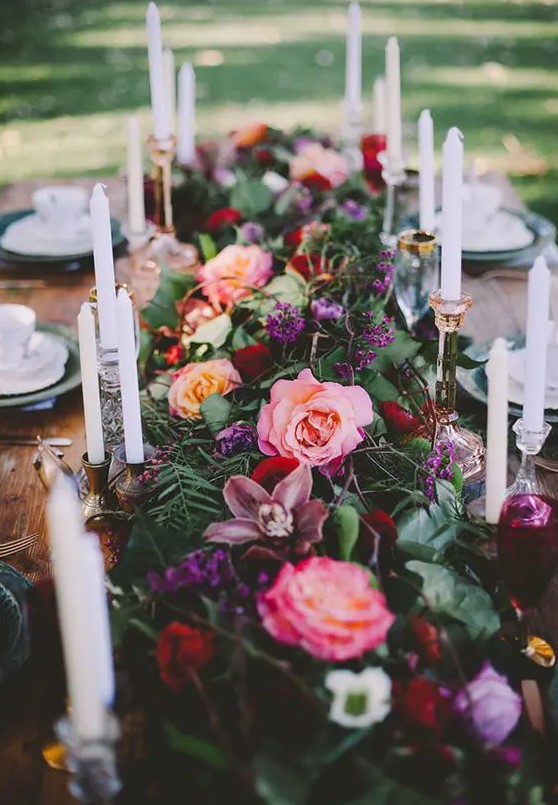 a moody floral table runner with blooms of different colors - [ink, mauve, purple and textural greenery dotted with candles