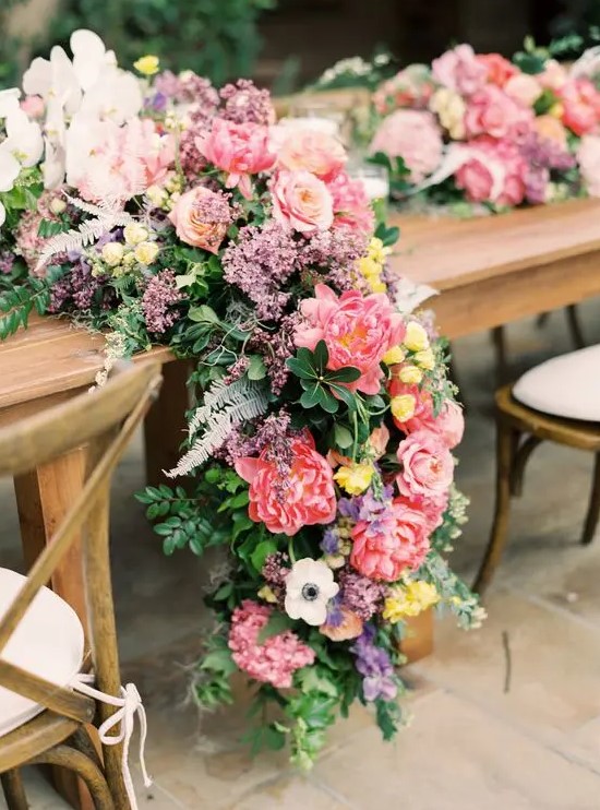 a super lush and colorful wedding table runner of pink roses and peonies, white anemones and purple flowers and textural and dimensional greenery