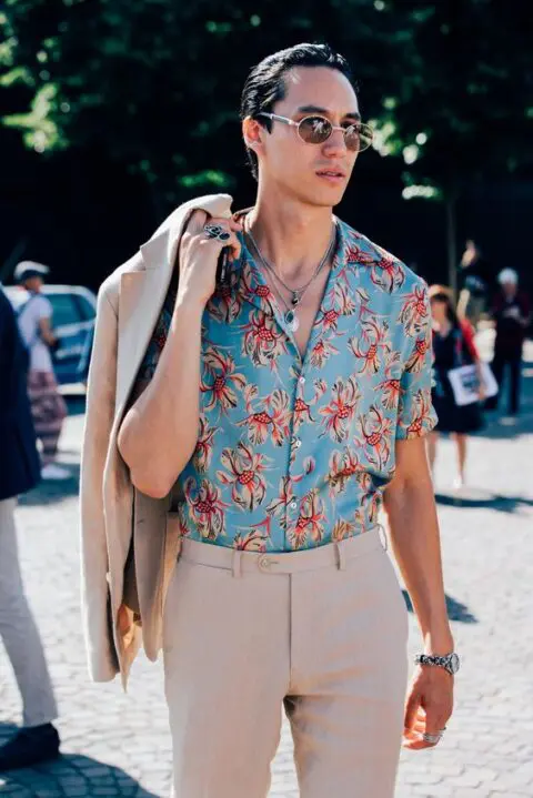 a bold tropical wedding guest look with a blue printed shirt, a tan linen suit and layered necklaces is cool and chic