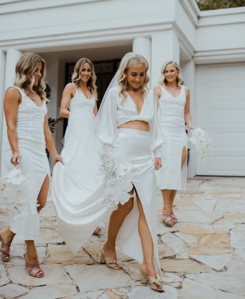 a bride wearing a neutral twisted crop top with puff sleeves and a maxi skirt with a slit, gold mules and a veil, bridesmaids rocking midi white dresses with slits and dusty pink mules