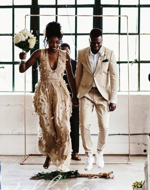 a bride wearing a tan floral applique wedding dress and a groom rocking a tan pantsuit, a white shirt and white sneakers