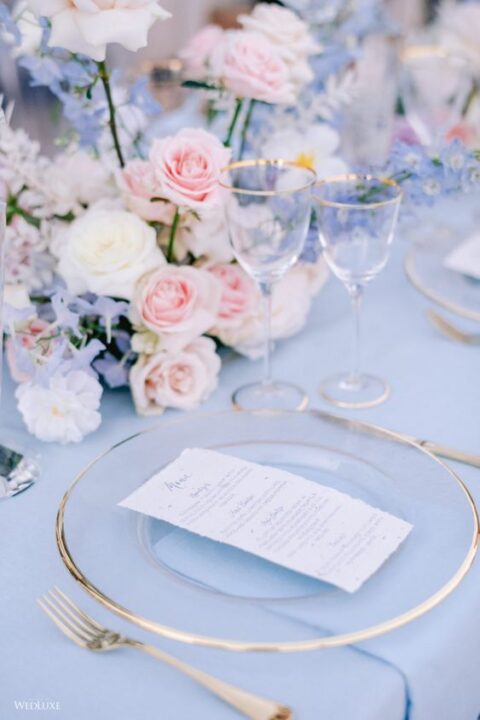 a beautiful pastel wedding tablescape with a blue tablecloth, pink, blue and white florals and clear plates with a gold rim
