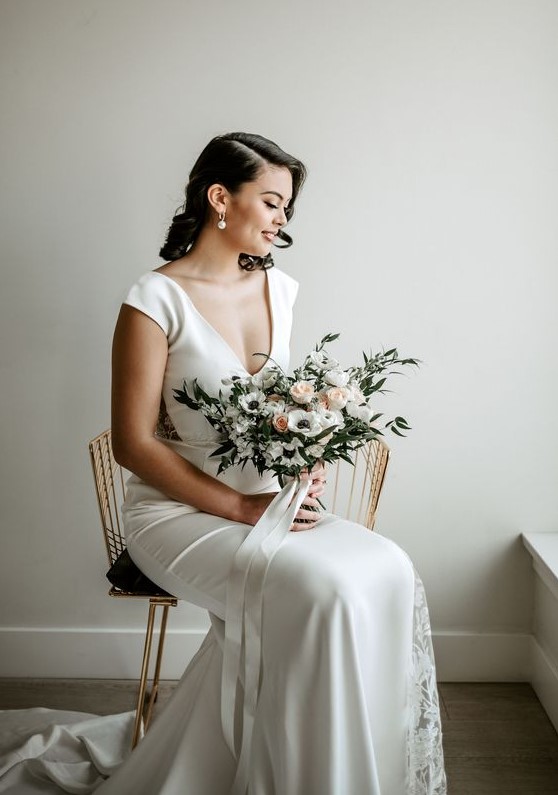 a modern glam bridal look with a plain fitting wedding dress with lace inserts, a deep neckline and cap sleeves plus statement pearl earrings