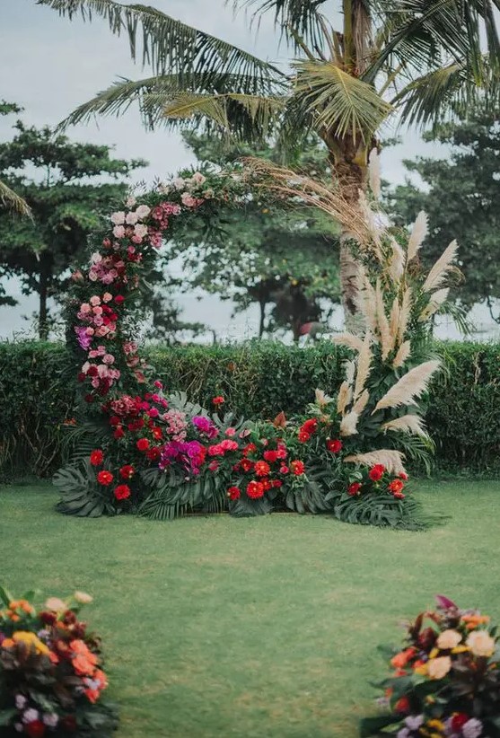 a lush round wedding arch with pampas grass, pink, blush, red and fuchsia blooms and tropical leaves