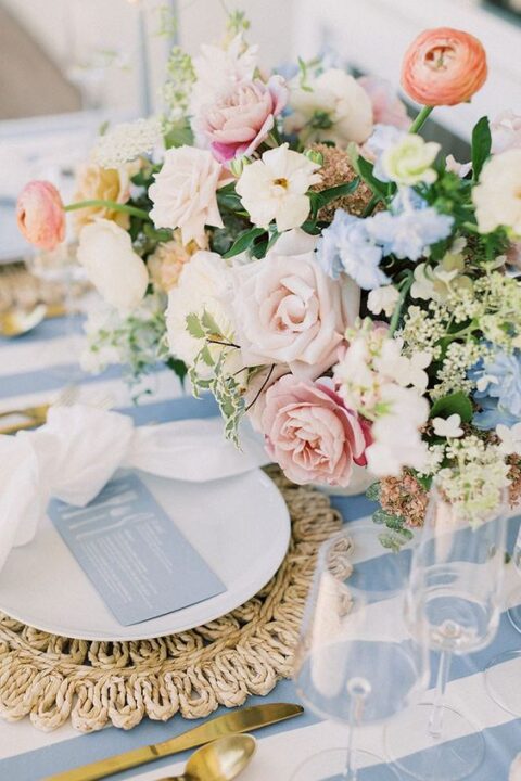 a lovely pastel wedding tablescape with a striped tablecloth, a woven placemat, neutral napkins, a gorgeous pastel floral centerpiece