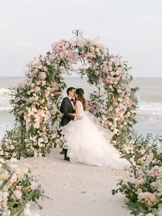 a beautiful and lush curved wedding arch covered with greenery, neutral and blush blooms and with a matching wedding aisle plus a sea view