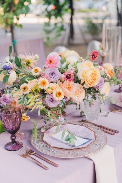 a lush pastel wedding tablescape with a lilac tablecloth, lilac, pink, peachy and purple blooms, purple glasses and printed placemats