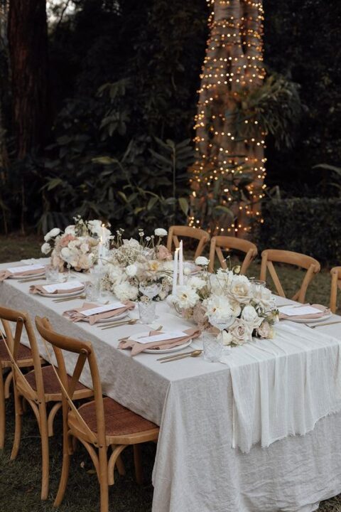 a chic and lovely neutral wedding tablescape with neutral linens, dusty pink napkins, white blooms and candles and gold cutlery