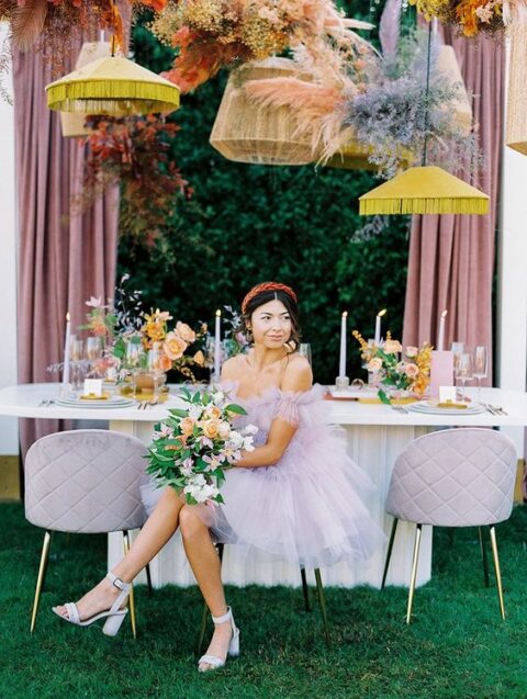 a pastel wedding reception space with yellow pendant lamps, pastel blooms and pampas grass, the bride wearing a lilac dress and shoes